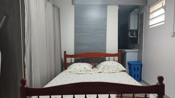 Ein kleiner, schmaler Raum mit einem Einzelbett, einem blauen Mülleimer und einem weißen Waschbecken in der Ecke.