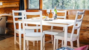 L’intérieur d’un chalet en bois, avec une table à manger dressée pour deux personnes, des chaises blanches et une bouteille de vin posée sur la table.