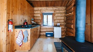 L’intérieur d’une cabane en rondins, avec un décor en bois, un coin cuisine comprenant un poêle, un réfrigérateur et un tonneau bleu.