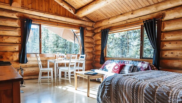 Une chambre dans une cabane en rondins, avec un plafond en bois, un lit recouvert d’une couverture à rayures, un bureau avec une chaise et une fenêtre avec des rideaux.