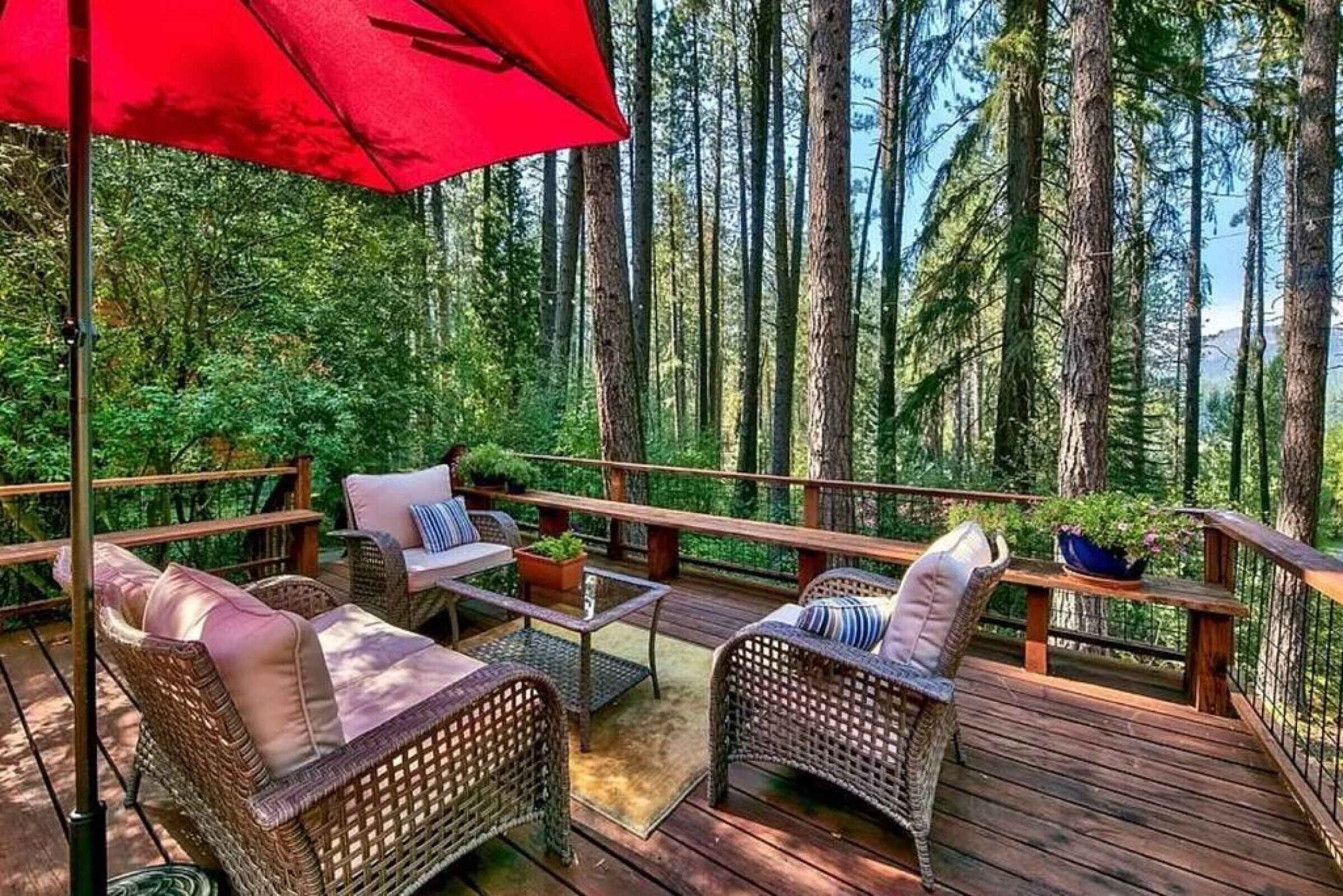 Eine Holzterrasse mit Korbmöbeln und einem roten Sonnenschirm, umgeben von hohen Bäumen.