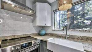 Una cocina moderna con armarios blancos, una estufa de acero inoxidable, horno integrado, encimera de granito, un lavabo blanco y un amplio ventanal con vistas a árboles.