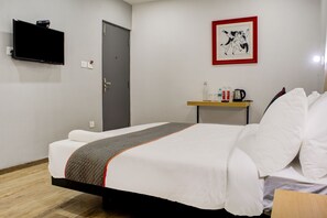 Habitación de hotel con cama, televisor montado en la pared, puerta, mesita con hervidor y botellas, y un cuadro enmarcado.