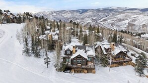 A log cabin with a stone foundation, surrounded by snow-covered trees and a ski slope.