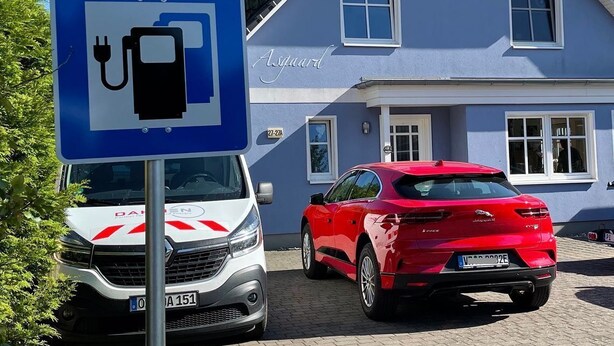 Ein blaues Schild mit einem Stecker-Symbol, das eine Ladestation kennzeichnet, ein weißer Lieferwagen und ein rotes Auto, geparkt vor einem Haus mit der Aufschrift 'Asgaard' an der Fassade.