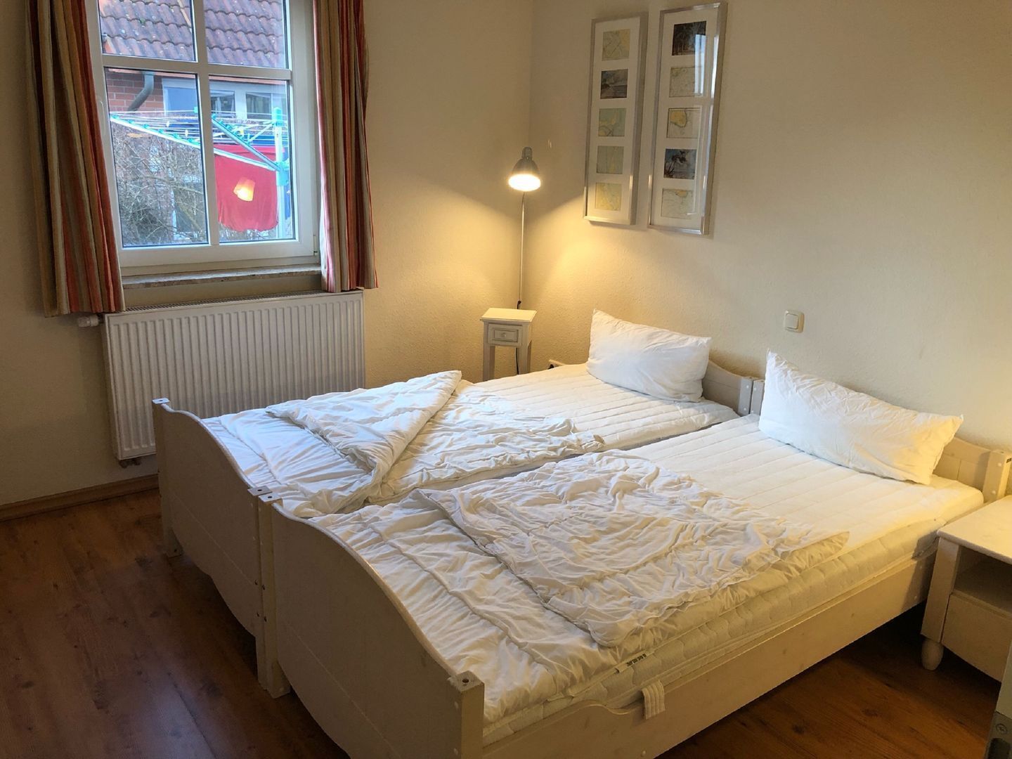 Une chambre avec deux lits, une fenêtre avec des rideaux, un radiateur, une table de chevet et des tableaux accrochés au mur.