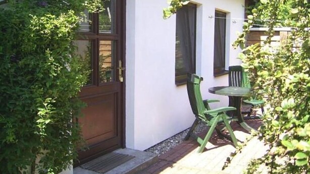 A white building with a wooden door and windows, a small outdoor seating area with a table and chairs, and a brick pathway leading to the entrance.