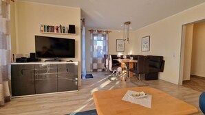 Una sala moderna con televisor, estanterías, comedor con mesa y sillas, y un sofá.