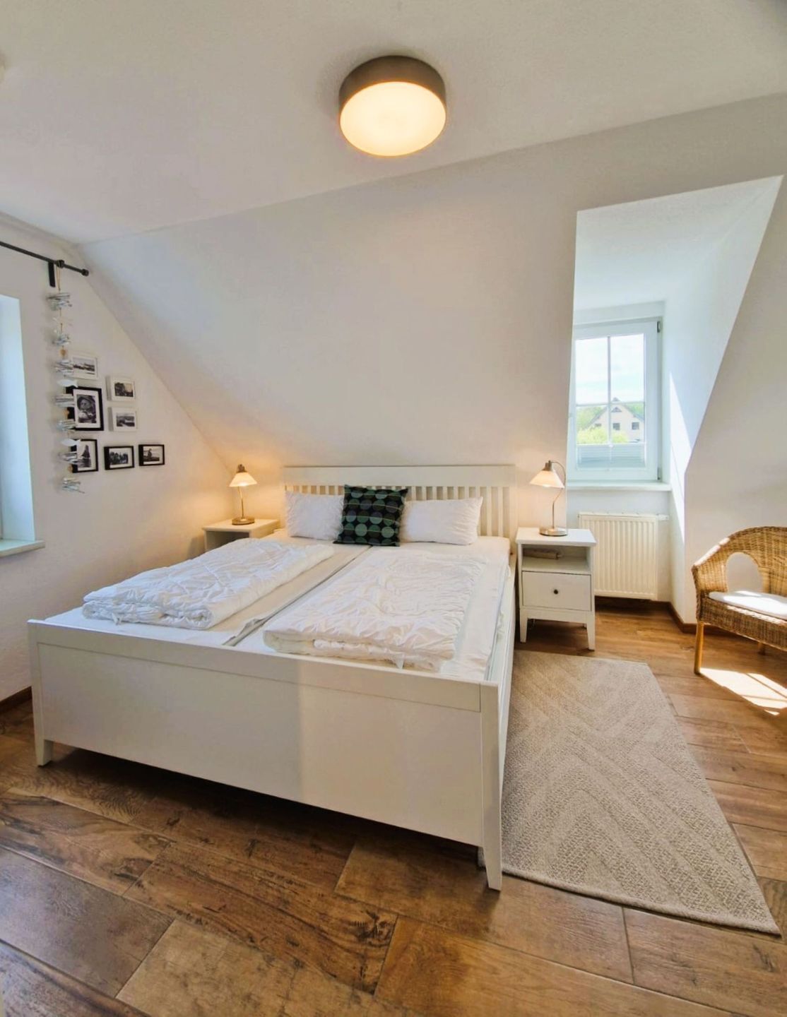 Une chambre à coucher avec un lit blanc, une table de chevet, une lampe, une chaise en osier, une fenêtre et un luminaire au plafond.