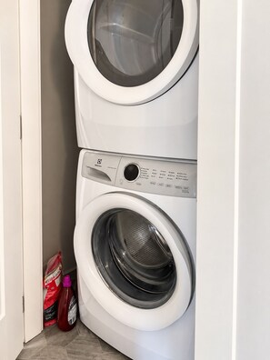 A front-loading washing machine with a control panel, a top-loading dryer, and cleaning supplies on the floor. - Zen Mountaire retreat, 1 bedroom, 1 bath, private parking (Langford)