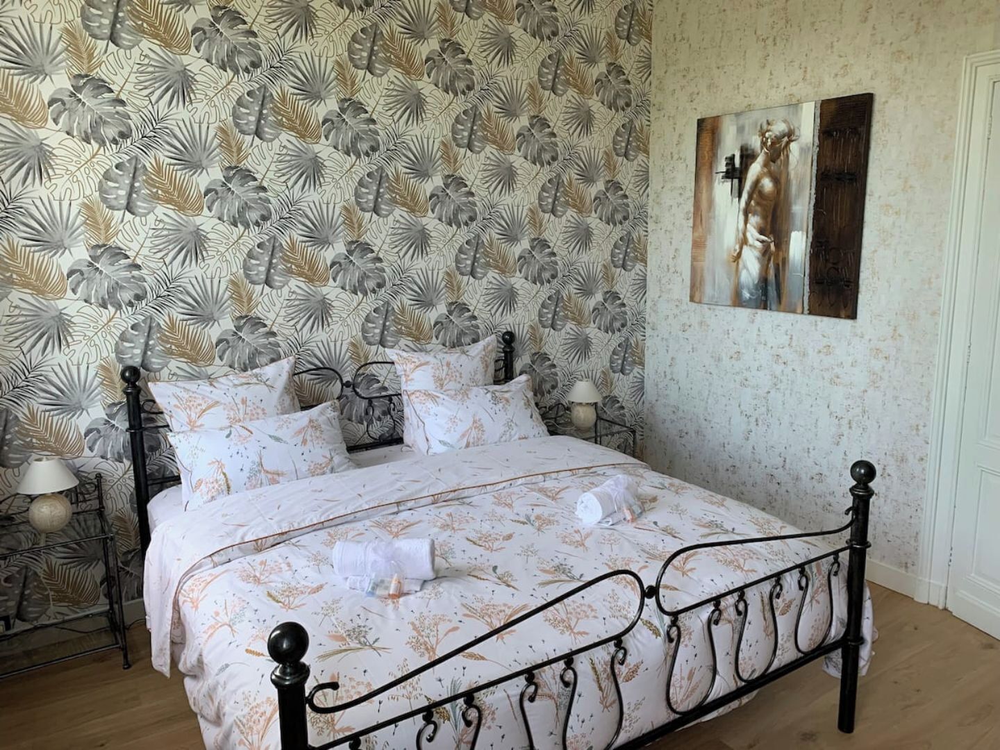 Quarto com papel de parede estampado com flores, uma cama com estrutura de metal e um quadro na parede.