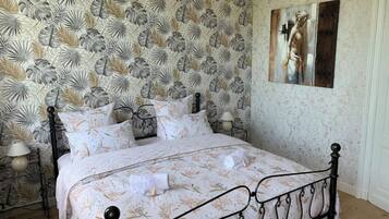 Quarto com papel de parede estampado com flores, uma cama com estrutura de metal e um quadro na parede.