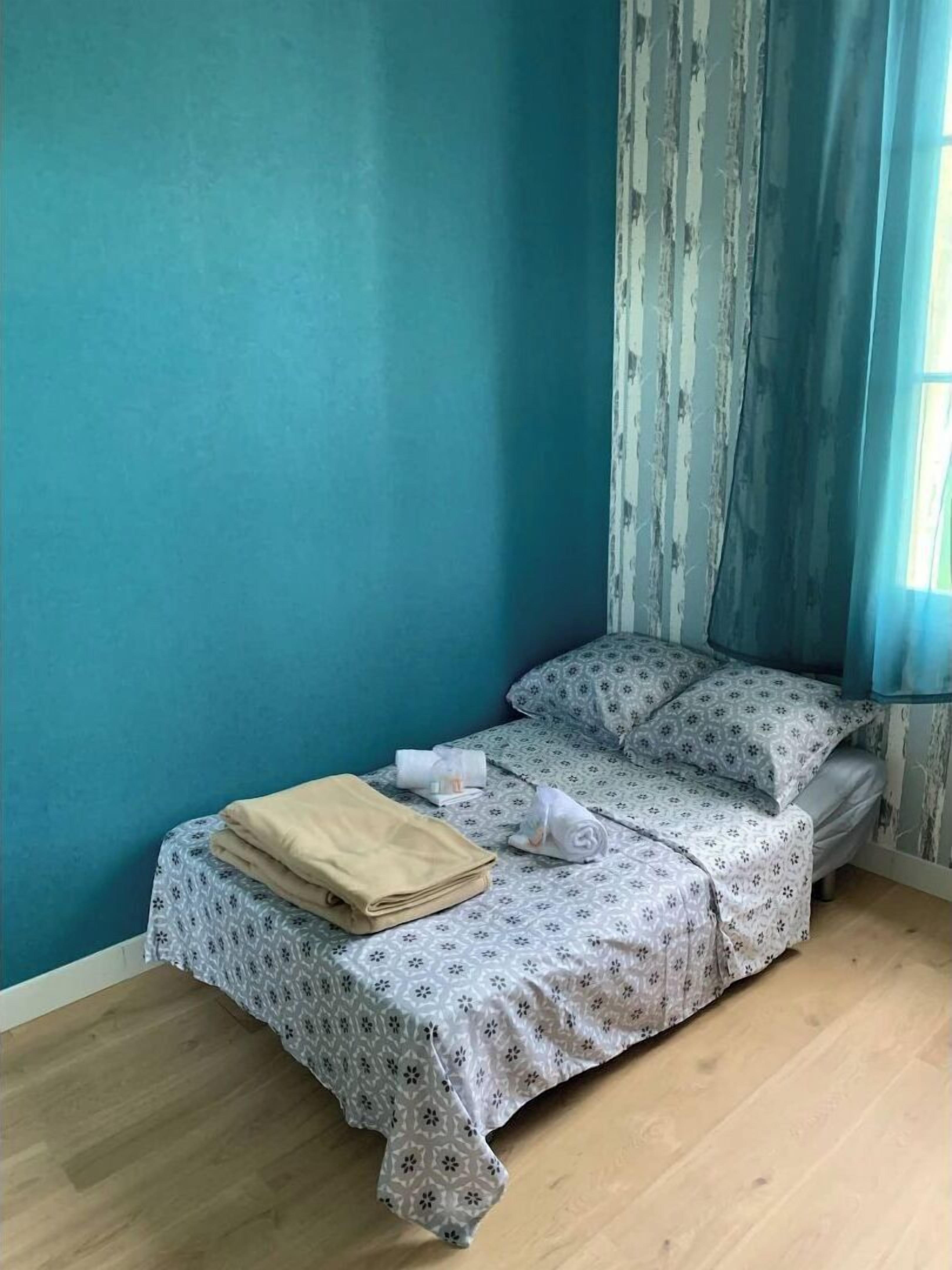 Ein kleines, ordentlich bezogenes Bett mit einem gemusterten Bettbezug und Kissen, einer gefalteten beigefarbenen Decke und einem aufgerollten weißen Handtuch.