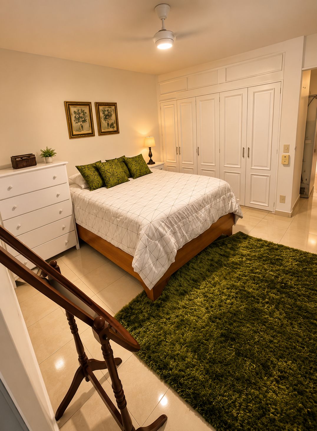 A bedroom with a bed, a dresser, a nightstand, a lamp, a ceiling fan, and a green rug.