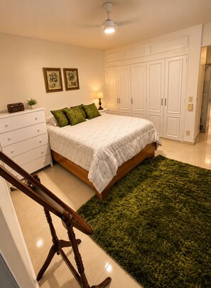 A bedroom with a bed, a dresser, a nightstand, a lamp, a ceiling fan, and a green rug. - Marants Lodge #2 (Puerto Plata)