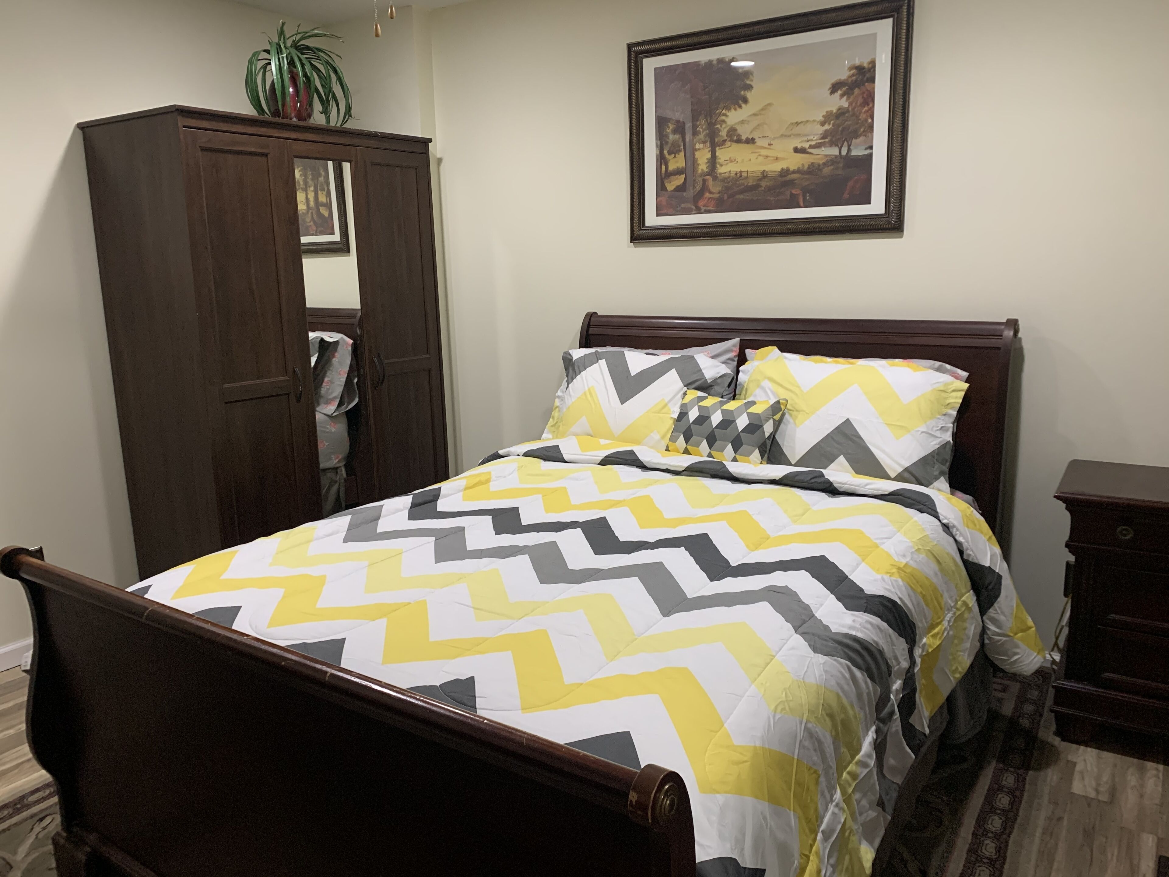 A bedroom with a bed, a wardrobe, a nightstand, a framed picture on the wall, and a potted plant.