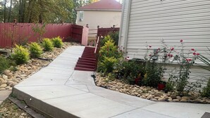 Un camino de entrada residencial con un sendero de concreto que lleva a una escalera de madera, rodeado por un jardín de rocas y diversas plantas en macetas.
