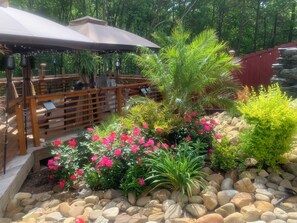 Un área de estar al aire libre con mesas y sillas de madera, rodeada de un jardín con diversas plantas y flores.