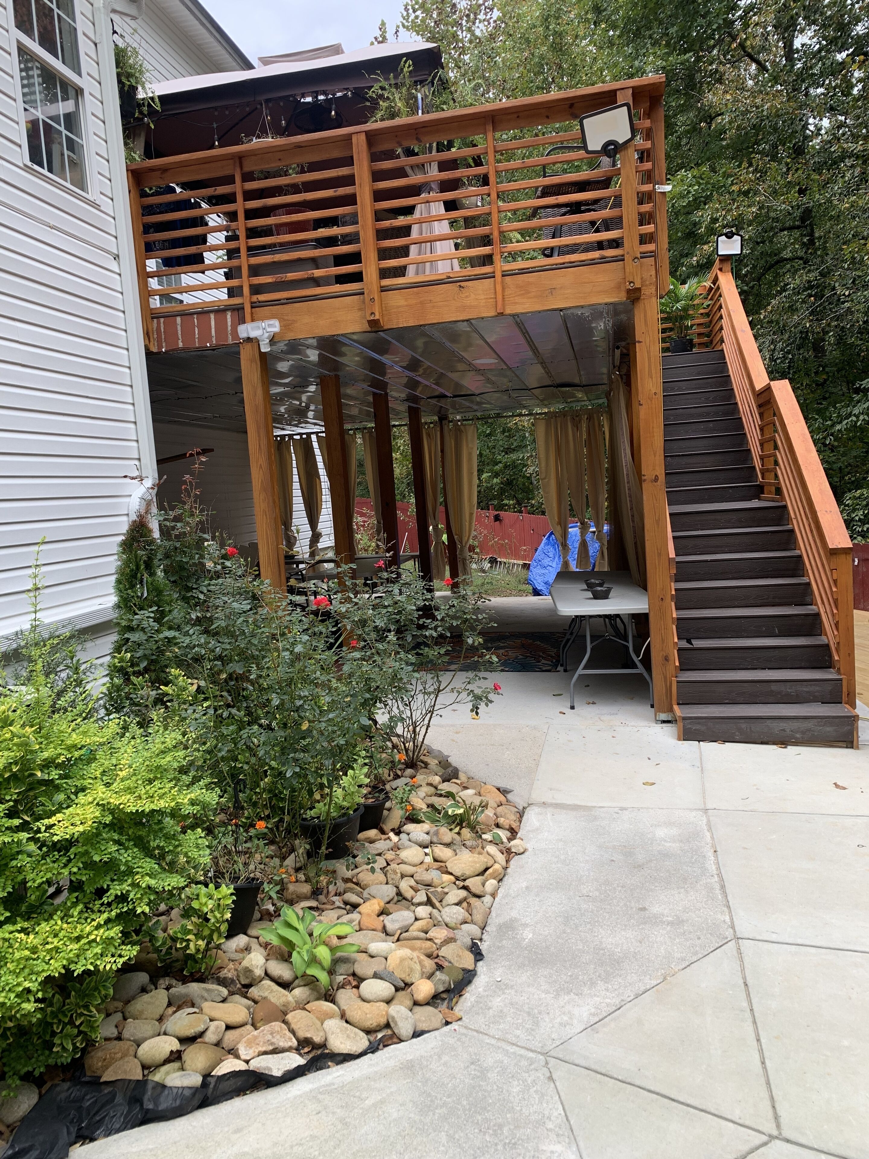 Un patio couvert avec une terrasse en bois, un chemin en pierre et un petit jardin agrémenté de diverses plantes et de rochers.