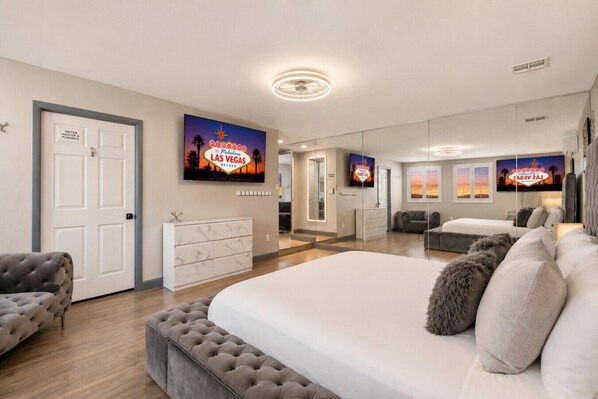 Une chambre spacieuse avec un grand lit, un canapé gris et un téléviseur diffusant une image d’un panneau de Las Vegas.