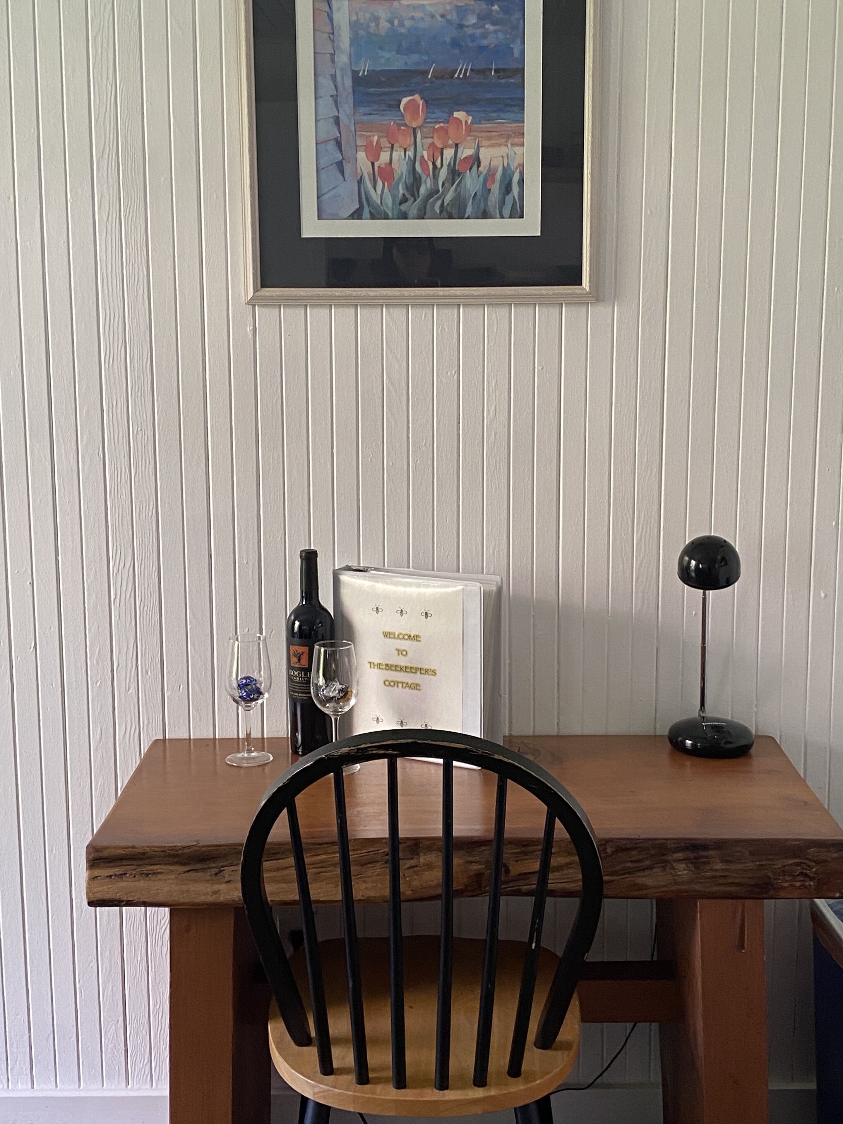 Una mesa de madera con un cuadro enmarco de un paisaje, una botella de vino, una copa de vino y un cartel de bienvenida.