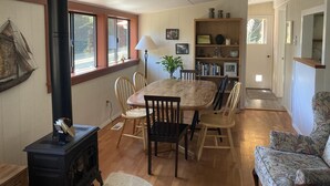 A cozy living room with a wooden table, chairs, a black wood stove, a bookshelf, and a patterned armchair.