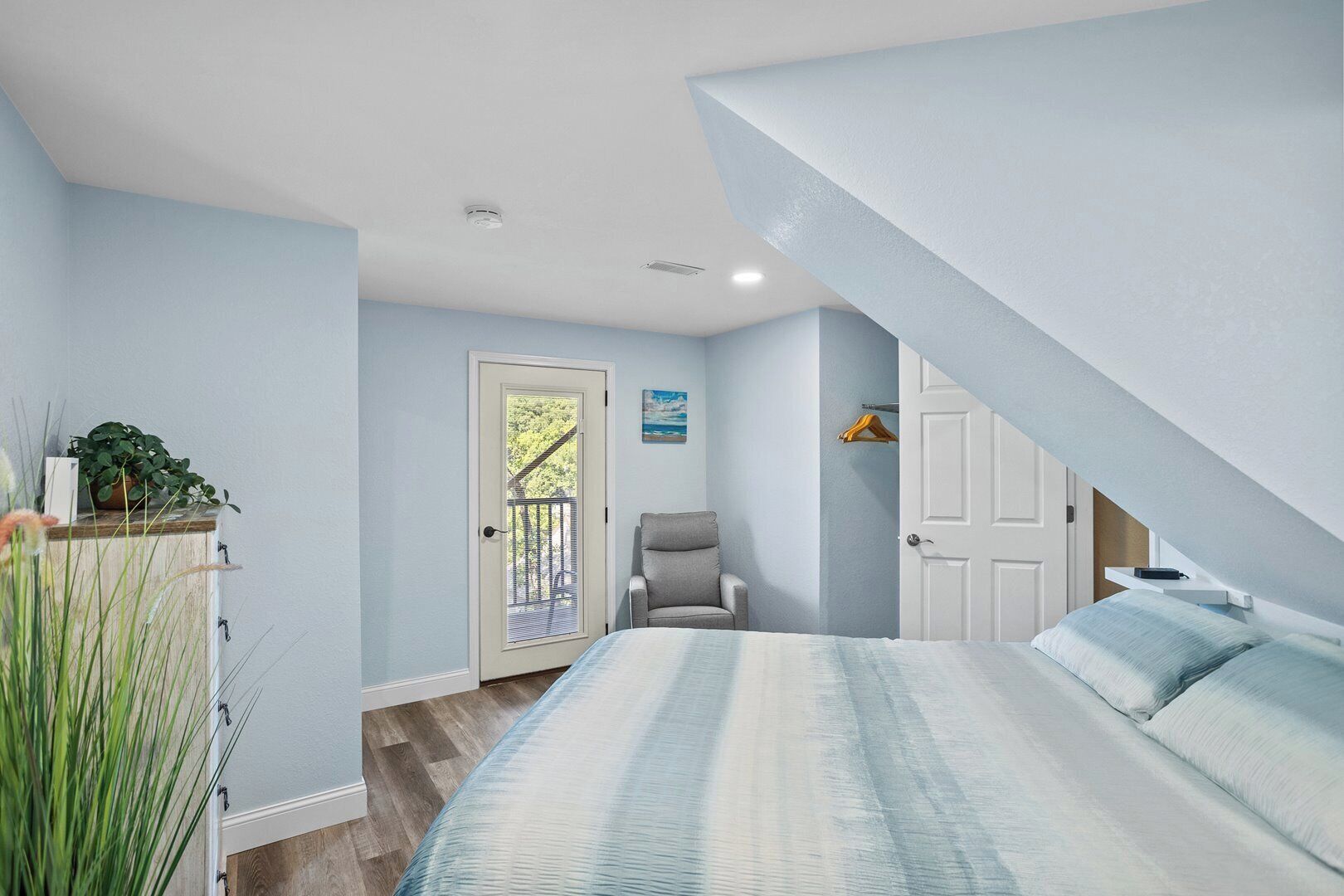 A bedroom with a bed, a chair, a door with a view, and a small shelf with a plant.