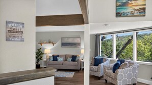 A spacious living room with a large window, a sofa, two patterned armchairs, a coffee table, and a decorative wall sign.