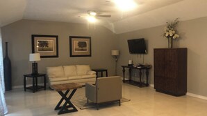 A living room with a high ceiling, a ceiling fan, a flat-screen TV, a sofa, a coffee table, a cabinet, and framed pictures on the wall. - Spacious 2300SF, Recently Rehabbed, Large Living (Crosby)