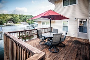 A wooden deck with a table and chairs, a red umbrella, and a view of a lake with boats.