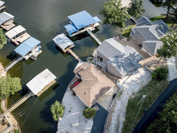 An aerial view of a lakeside property with docked boats, a house with a sloped roof, and a small dock extending into the water.