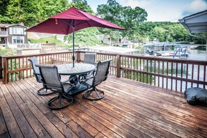 A wooden deck with a round table and chairs, a red umbrella, and a view of a lake with boats.