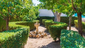 Un jardín bien cuidado con setos ordenados, una fuente de piedra en el centro y diversas especies de árboles.