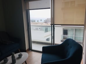 Una habitación con un sillón azul, una mesita con objetos decorativos y vista a un edificio a través de un ventanal grande.