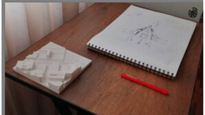 Et træbord med en hvid arkitektonisk model, en skitsebog og en rød pen.