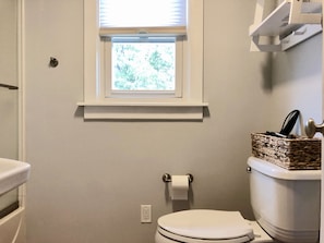 A bathroom with a toilet, a window, a shelf above the toilet, and a basket on the right.