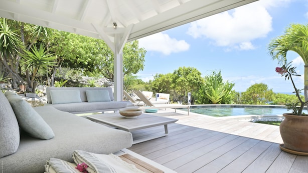 A wooden deck with a sofa and cushions, a table with a bowl, and a view of a swimming pool and greenery.