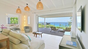 A modern living room with a sofa, chairs, a coffee table, and a TV. There is a balcony with a view of the ocean.