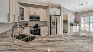 A modern kitchen with white cabinetry, stainless steel appliances, a marble countertop, and a view of the outdoors through large windows. - Naples- Briarwood Estates all new 4BR/3 BA, sleeps 10, vacation rental (Naples)