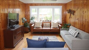 A living room with wooden walls, a brown leather sofa, a grey couch, a television on a wooden cabinet, and large windows with a view of the outside. - Blue House in Smallwood NY, Year round, 2-bed in Catskill Mts (Monticello)