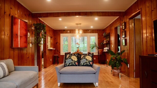 A cozy room with wooden walls, a grey sofa, a wooden floor, and a window with a view of trees. - Blue House in Smallwood NY, Year round, 2-bed in Catskill Mts (Monticello)