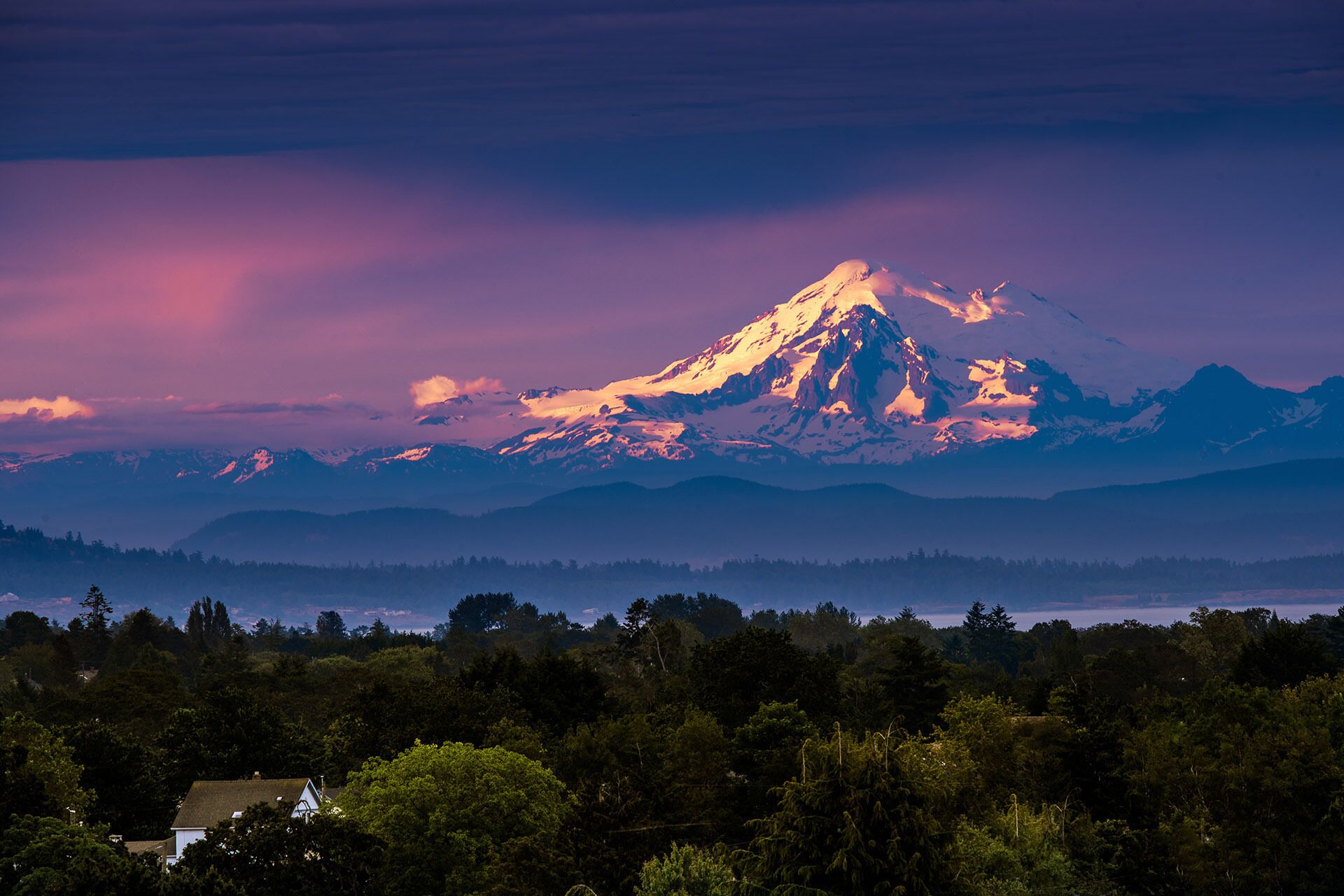 A mountain with snow-capped peaks and a forested valley below.