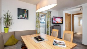 Ein Zimmer mit einem Holzesstisch, Stühlen, einer Couch, einer Topfpflanze, einem Fernseher und einer Tür zu einem anderen Raum.