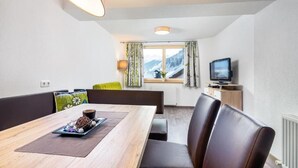 Een moderne eetruimte met een houten tafel, zwarte lederen stoelen, een televisie op een meubel en een raam met uitzicht op besneeuwde bergen.
