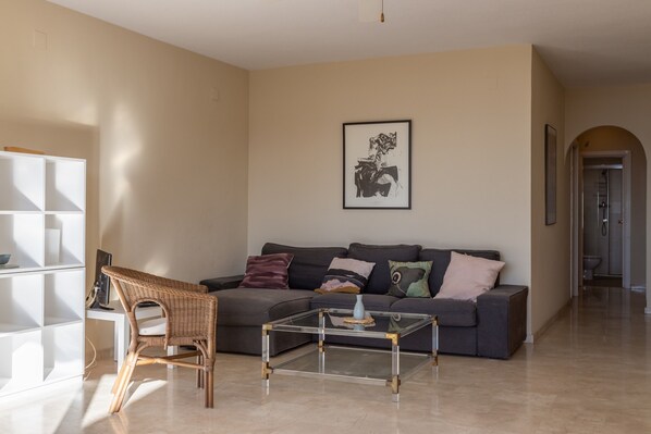 Una sala de estar con un sofá, una mesa de centro de vidrio, un sillón de mimbre y una obra de arte enmarcada en la pared.