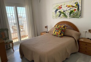A bedroom with a bed, a nightstand, a chair, a window with curtains, and a wall painting of parrots.