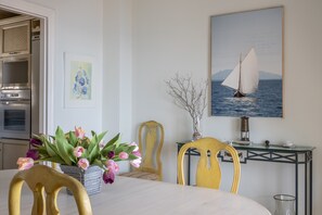 Un coin repas avec une table, des chaises, un vase de fleurs, un tableau représentant un voilier, et une cuisine en arrière-plan.