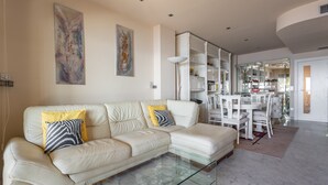 A modern living room with a white leather sectional sofa, a glass coffee table, and a dining area with white chairs and a table.