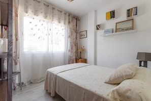 A bedroom with a bed, a nightstand, a lamp, a window with curtains, and wall-mounted shelves with books and pictures.