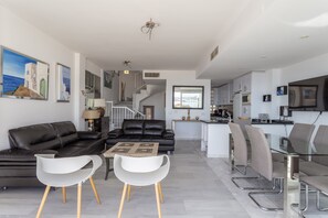 A modern living room with a black leather sofa, white chairs, a glass coffee table, and a staircase leading to a kitchen.